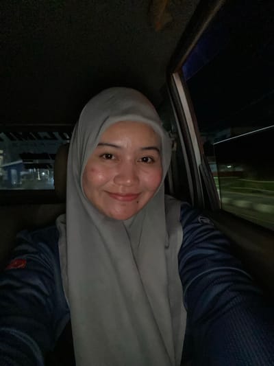 The media file shows a close-up selfie of a woman wearing a light gray hijab and a dark blue shirt. She is looking at the camera with a slight smile, her face illuminated by soft lighting. The background is dark, suggesting an indoor setting at night, possibly in a car due to the visible car interior elements like the window frame and what appears to be a sunroof. Her shirt has a visible logo on the sleeve, which is red and white. The image is oriented sideways. The location context provided is Jawa, Indonesia. There is no visible text in the image itself.