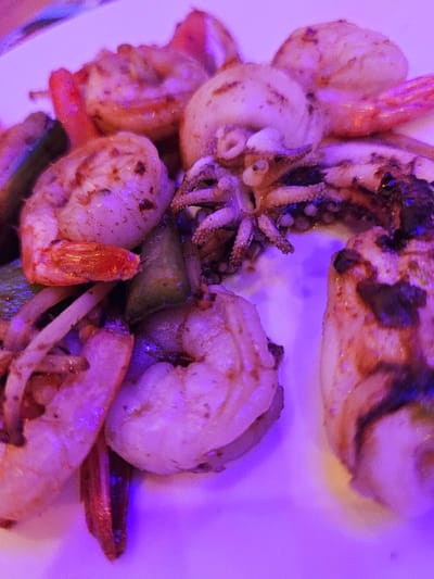 A white plate, under a predominant magenta-purple illumination, displays a cooked seafood and vegetable arrangement. The primary subjects are approximately six to eight shrimp, peeled, with some tails remaining, showing orange-red coloration and light charring or seasoning marks. Interspersed among the shrimp are multiple pieces of cooked cephalopod; one distinct piece features a cluster of tentacles, while other larger segments exhibit charring and cross-sections consistent with grilled squid or octopus. Scattered green vegetable fragments, resembling bell peppers or zucchini, and thin white slices, possibly onion, are mixed with the seafood. The items are stationary on the plate, indicating a served meal. This scene is located in Audun-le-Tiche, France.