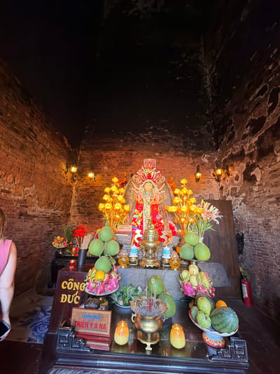 This image depicts an indoor shrine, likely in a religious or cultural site in Tuy Hoa Ward, Vietnam. The shrine is set within a room with rustic brick walls and a dark ceiling. Dominating the scene is a central altar displaying a statue of a deity adorned in red and gold. The altar is laden with offerings, including an abundance of green pomelos, other fruits like dragon fruit, mangoes, watermelons, and oranges, arranged on platters. Several lit candles and incense sticks in a brass urn are also present, suggesting active worship. Decorative elements include golden artificial flowers and a brass urn. Two wall-mounted lamps cast a warm glow on the scene. In the foreground, to the left, a person wearing a pink top is partially visible, facing away from the camera and holding a smartphone. A fire extinguisher is mounted on the wall to the right, behind the altar. Signs with Vietnamese text are placed on the altar. The overall atmosphere is one of reverence and tradition.