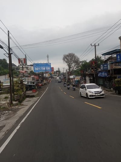 The image shows a street in an urban area. The road is paved and has a yellow line marking the lanes. There are several vehicles on the road, including a white car, motorcycles, and a truck. There are buildings on both sides of the road, including shops and restaurants. There are also utility poles and overhead wires. There is a KFC sign in the distance. The sky is overcast. It is unlikely this image is from Taunggyi, Myanmar, as the license plates are not consistent with plates issued in Myanmar. The image most likely depicts a street scene in Indonesia, as the license plate on the white car is an Indonesian plate. The street appears to be in a commercial or residential area.