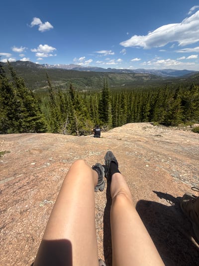 A first-person perspective shot captures a person's legs and feet resting on a large, pink granite rock formation. The person is wearing black hiking shoes and black socks. In the background, a vast expanse of dense green pine forest stretches towards a range of distant mountains under a bright blue sky with scattered white clouds. A lone figure is visible in the middle ground, sitting on the forest edge. The scene appears to be in a natural, mountainous environment, possibly in or near Estes Park, Colorado, given the characteristic rocky terrain and coniferous forests often found in that region.