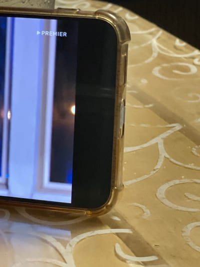 A close-up, side-view photograph shows a smartphone screen displaying the word "PREMIER" with a play icon to its left, superimposed on a slightly blurred background. The phone is positioned on a table with a light yellow textured surface featuring white swirling patterns. The phone itself has a gold-colored edge and a clear protective case. The lighting in the image suggests an indoor setting, possibly at night, given the dim reflections on the screen and the overall ambiance. The scene does not provide any specific geographical markers to indicate its location within the city of Mirnyy, Russia.