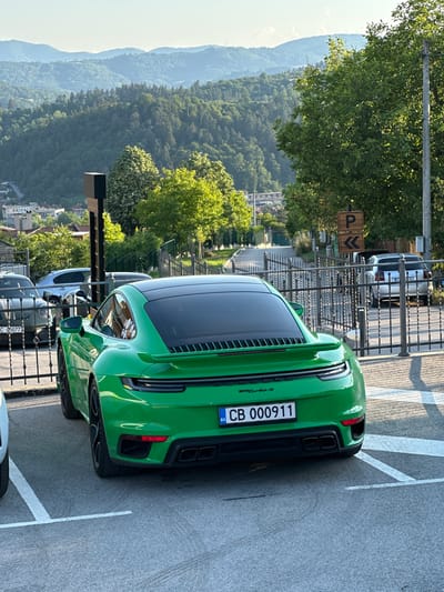 A bright green Porsche 911 Turbo S is parked in an outdoor parking lot. The car is the primary subject, showcasing its rear end with the license plate visible. The license plate reads "CB 000911" and has a blue EU strip. The car is positioned on asphalt marked with white parking lines.

In the background, a metal fence separates the parking lot from a scenic view. This view includes lush green trees covering rolling hills and mountains, suggesting a natural, possibly mountainous, landscape. There are also buildings visible in the distance, hinting at a town or village nestled within the landscape. The sky is bright and clear, indicating daytime with fair weather.

Two parking signs are visible on a pole near the fence. One sign is square with a brown background and a white 'P' symbol, indicating parking. The other sign is rectangular with a brown background and white arrows pointing left and down, possibly indicating directions or restrictions related to parking.

Other cars are partially visible in the parking lot, including a silver car and a white car further back. The overall scene conveys a sense of a well-maintained parking area in a picturesque location. The image is oriented vertically, as if taken from a higher vantage point looking down or from a vehicle moving past.