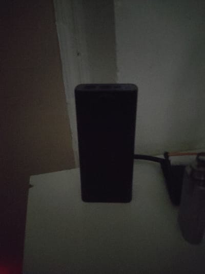 A black rectangular power bank sits on a white surface. The power bank has two visible ports at the top. To the right of the power bank, a black cable connects to a device that is mostly out of frame. The background consists of a textured wall. The lighting is dim, casting shadows and obscuring details. The scene appears to be indoors, on a flat surface. There is no indication of the city of Кипчок, Tajikistan.