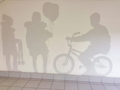 The image shows a mural on a light-colored wall depicting silhouettes of people and objects. On the left, there are two children, one holding what appears to be a bouquet and two balloons. Next to them, a child is depicted with what looks like a jacket with a furry trim. To the right, a silhouette of a person is shown riding a bicycle. The mural is situated above a tiled baseboard and a tiled floor. The overall impression is a playful and whimsical scene. While the prompt specifies the location as Kurgan, Russia, this information cannot be visually verified from the image itself.