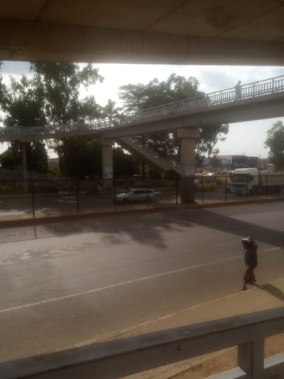 The image shows an elevated pedestrian walkway above a road in Unguwan Dosa, Nigeria. The walkway is supported by concrete pillars and has railings on both sides. There are a few people walking on the walkway. Below the walkway, a road is visible with several vehicles, including cars, a yellow minivan, and a white truck. A person is walking on the side of the road in the foreground, carrying a load on their head and back. Trees and buildings can be seen in the background. The lighting suggests it is daytime, possibly morning or afternoon, with a mix of sunlight and shadows. The sky appears overcast. The overall scene depicts an urban environment with traffic and pedestrian infrastructure. No visible text is discernible in the image.
