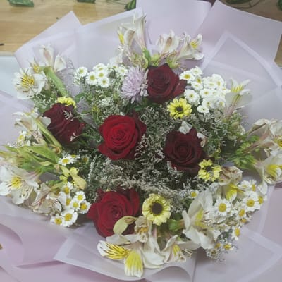 A bouquet of flowers is displayed, wrapped in pale pink and white paper. The bouquet is composed of several red roses, white alstroemeria with yellow markings, small white daisies with yellow centers, and some delicate white filler flowers. A pale purple chrysanthemum is also visible. The flowers are arranged in a rounded shape. The background appears to be a wooden surface, possibly a table. The image is taken from a slightly elevated perspective. The location is specified as Urgench, Uzbekistan, suggesting this is likely a scene within the city. There are no people or actions depicted in the image.