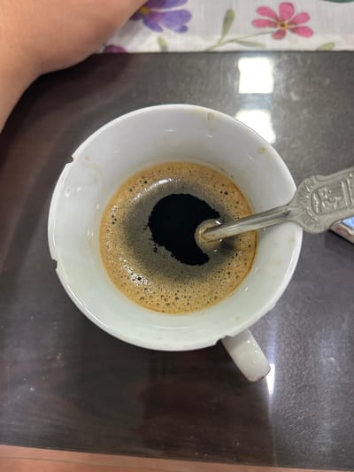The image shows a close-up, overhead view of a white ceramic mug filled with dark coffee and a layer of crema. A metal spoon with an embossed pattern is partially submerged in the coffee. The mug sits on a dark, reflective surface, likely a table. In the background, a colorful tablecloth with floral patterns is visible, along with a person's arm. The lighting suggests an indoor setting. There is no indication of the city of Dulovo, Bulgaria, in the image itself.