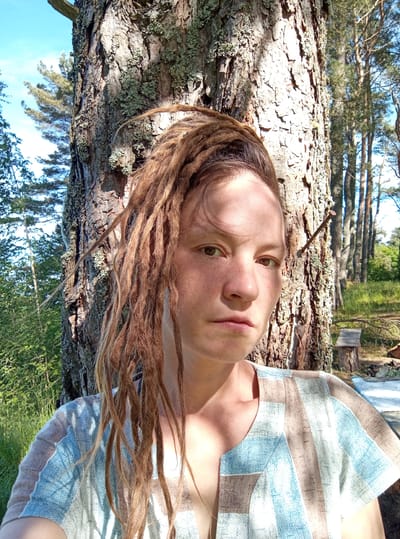 The image depicts an adult individual with light brown dreadlocks, pulled back from the face, standing outdoors in a natural environment. The person has fair skin with visible freckles on the nose and forehead, and a neutral facial expression, looking directly forward. They are wearing a short-sleeved top featuring a geometric pattern of light blue, tan, and white blocks.

The individual is positioned against the rough bark of a large tree trunk, which dominates the left side of the frame and extends upwards. The tree bark is covered with patches of light green lichen. To the right of the person and the main tree, a dense stand of tall, slender conifer trees forms a forest background under a clear blue sky. Sunlight casts partial shadows across the person's face and onto their clothing. A rusted metal nail is partially embedded in the tree trunk to the person's right, angled upwards. In the far right background, a rustic bench made from a cut log is visible on the grassy ground, with other fallen tree debris nearby. The scene is situated in the city of Podborov'e, Russia.