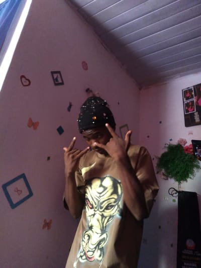 The image depicts a young person wearing a black beanie and a tan t-shirt with a graphic design of a face on it. The person is standing in what appears to be a room, with a light-colored ceiling made of horizontal planks. The walls are painted a light pinkish-purple and are decorated with various stickers and a framed picture. Some of the stickers are in the shape of hearts and butterflies, while others are small, abstract shapes. There is also a framed picture of what appears to be cartoon characters. In the background, a green plant in a pot is visible, along with a black object that could be a bag or a speaker. A framed picture of two young women in pink tops is hanging on the wall. The lighting in the room is somewhat dim, with a strong light source coming from the left side of the image, casting shadows. The overall impression is that of a personal space, possibly a bedroom, decorated with youthful and whimsical elements. The location being Kassa Market, Nigeria, suggests an urban setting within that specific market area.