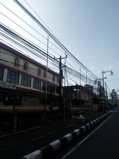 The image presents a street scene in Nhot Chang, Laos, characterized by numerous overhead power lines dominating the sky. A multi-story building with a cream and maroon facade is visible on the left side of the frame, with dark-tinted windows and signage in a local language. A chain-link fence runs along the base of the building.

Further down the street, there is a building under construction with exposed wooden framing. Several utility poles are supporting the tangle of wires. A row of decorative street lamps with birdcage-like enclosures lines the right side of the street.

The road surface is dark asphalt with a painted white line dividing it. A curb painted with alternating black and white stripes borders the road. A few people are visible in the distance on the right side of the street. The overall lighting suggests early morning or late afternoon.