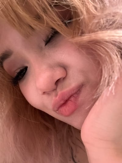 A close-up, low-angle, indoor shot shows the face of a young woman with blonde hair. She is looking down and to the left, with her left eye closed and her right eye partially open. Her lips are slightly parted in a pout. The background is blurred and appears to be a dimly lit room, possibly a bedroom, with warm lighting. The image was taken in Anadyr, Russia.