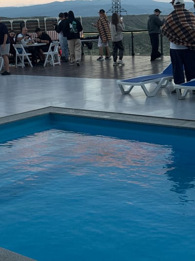 The image depicts a group of people gathered on a wooden deck next to a swimming pool. The perspective is slightly angled, and the image is oriented vertically.
In the foreground, a portion of the swimming pool is visible, filled with clear blue water. The surface of the water is rippled, reflecting some of the surrounding environment, including what appear to be warm-toned colors. A grey, textured edge surrounds the pool.
To the left of the pool is a wooden deck, and beyond that, a tiled area with white lounge chairs. Several people are scattered across the deck and the tiled area. One person in the upper left is wearing a plaid shirt and dark pants. Further down the deck, a few individuals appear to be standing and looking out, while others are seated at white tables and chairs. Some of these individuals are dressed casually in t-shirts and pants.
In the background, a railing separates the deck area from a sloping landscape covered in vegetation. Mountains are visible in the distance, suggesting a hilly or mountainous terrain. A large metal tower, possibly a power line pylon, stands on the left side of the frame.
The lighting suggests it is either early morning or late afternoon, given the soft light and potential for reflections. The sky appears overcast or hazy. The overall mood seems relaxed and social, with people enjoying a gathering by the pool in what appears to be a scenic outdoor location. The location context provided is Mukhatskaro, Georgia. There is no visible text in the image.