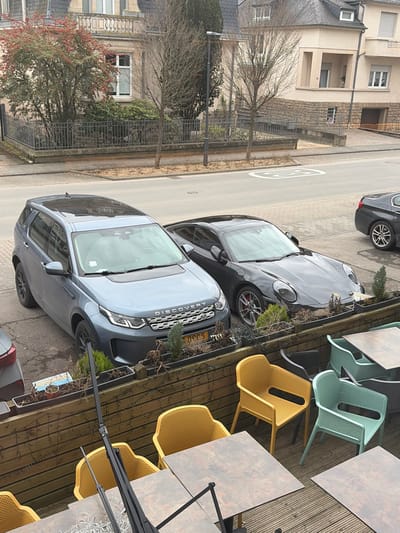 This image depicts a static street scene in Walferdange, Luxembourg, observed from an elevated position, likely a cafe or restaurant terrace.
In the foreground, a multi-colored outdoor dining area features wooden decking, several dark-topped tables with black bases, and numerous plastic chairs in yellow, light green, and dark grey. A rectangular wooden planter box, containing mostly dormant or dead plants interspersed with small evergreen shrubs, runs along the terrace's edge. A black metal support structure, possibly for an awning, extends upwards on the left.
Immediately beyond the terrace, on a paved surface, a grey Land Rover Discovery SUV (license plate D 09 C5, Luxembourgish registration) is parked facing slightly left. Behind it, a dark grey or black Porsche 911 coupe, identifiable by its distinctive headlights and red brake calipers, is parked. The rear section and a single wheel of a third dark-colored vehicle, possibly a BMW, are visible on the far right, further down the street.
The vehicles are parked alongside a narrow asphalt street that features a prominent white "30" speed limit marking painted on its surface. A paved sidewalk runs parallel to the street.
In the background, across the street, stand several multi-story buildings. An older structure on the left features a light-colored facade with intricate window details and a prominent bush adorned with red berries in front of a metal fence. To its right, a more contemporary building with a beige/cream facade and a sloped roof is visible. Bare deciduous trees, along with some evergreen shrubs, line the street and properties. A single streetlamp is visible on the left side of the street. The sky is overcast, indicating a cloudy day. No individuals are present in the image.