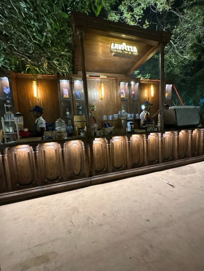 An outdoor Lavazza coffee stand is nestled amidst lush greenery. Its wooden structure features illuminated shelves displaying various items. A person wearing a white shirt and blue cap stands behind the counter. The stand has a rustic, wooden aesthetic, accented by warm lighting. A concrete or stone surface abuts the structure, possibly a sidewalk or patio.