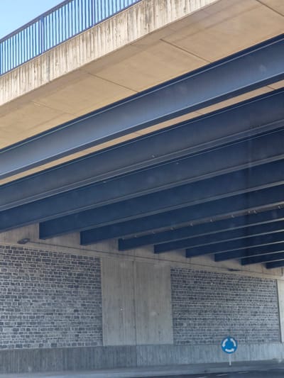 The image shows an underpass or tunnel in Dudelange, Luxembourg. The overhead structure is composed of concrete beams and a metal railing, with a sky visible above. Beneath this, dark blue metal beams are layered, supporting the upper level. The walls of the underpass are constructed from grey brick, with sections of plain concrete. A circular blue traffic sign indicating a roundabout is visible on the right side of the image, placed on a white pole near the road. The road surface appears to be asphalt, with white lane markings. The scene is a typical urban infrastructure element, designed to allow traffic to pass under a higher structure.