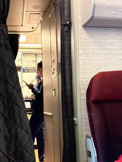 The image appears to be taken inside a passenger train or bus, looking down the aisle. A female employee in a dark blue uniform is holding a telephone handset and talking. She stands in front of white storage cabinets, likely containing food or supplies. Above is the luggage rack. A burgundy passenger seat is visible at the top of the frame, along with wall paneling. The location, Pudahuel, Chile, is assumed to be where the train is traveling or based; however, this detail is not apparent from the image itself.