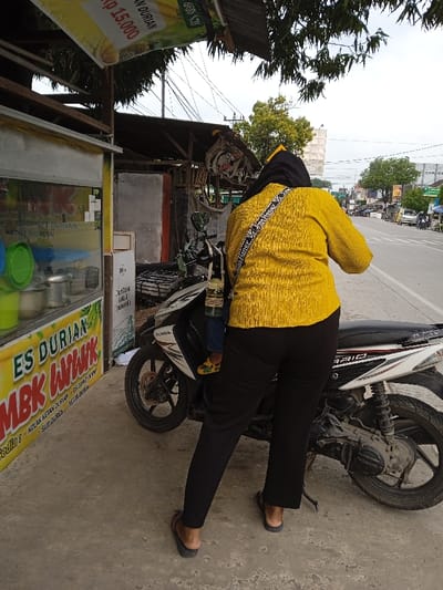 The image displays a roadside scene in Sarimulyo, Indonesia. An adult, viewed from the back, occupies the central foreground. This individual wears a yellow textured long-sleeved top, black trousers, and black flip-flops. A black head covering is visible, and a black and white patterned shoulder bag, featuring "Miss Honey" text, crosses their back diagonally. They stand on a concrete surface next to a white and black Honda Vario scooter. A small child, wearing blue pants and yellow sandals, is seated on the scooter's front portion, facing away from the camera. A transparent bottle hangs from the scooter's handlebars near the child. To the immediate left, a roadside food stall is visible, featuring a prominent yellow sign with red and black text reading "ES DURIAN MBK WIWIK," along with other menu items like "ENDUWET DURIAN" and "KETAN DURIAN," and pricing, including "Rp 15.000." The stall structure includes a glass display case containing green-lidded containers and metal pots. In the background, a two-lane asphalt road with a white solid line is visible, stretching into the distance where more buildings, stalls, and green trees are present under a bright sky with overhead utility lines.