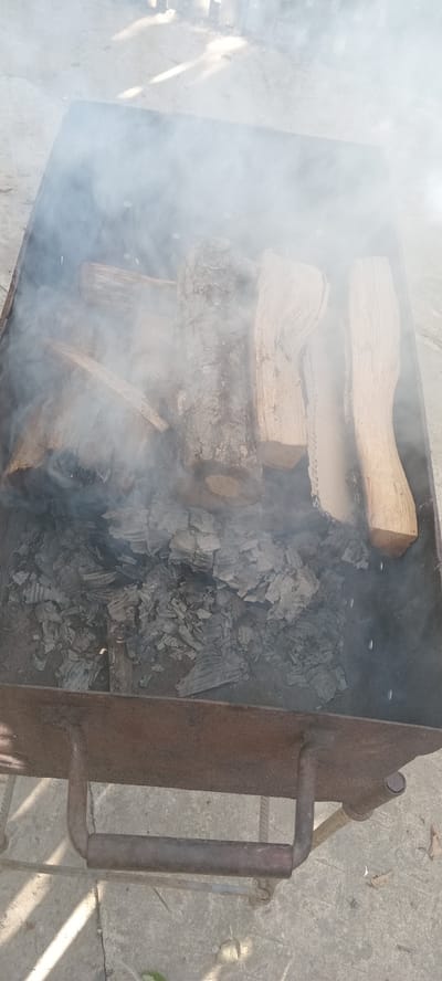 A top-down view of a rusty metal barbecue grill filled with partially burned logs and ashes. Smoke billows up from the grill, obscuring some of the contents. The grill is positioned outdoors on a concrete surface, likely in the city of Koviahy, Ukraine, as indicated by the context. The surrounding environment appears to be a casual outdoor setting, possibly a backyard or a park. The lighting suggests it is daytime, with shadows visible on the ground. There are no people or animals present in the image.