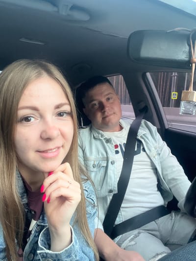The image shows two individuals, a woman and a man, in the front seats of a car. The woman is in the foreground, smiling at the camera, with her hand near her chin, adorned with bright pink nail polish and a ring. She is wearing a denim jacket over a dark shirt. The man is to her right, also looking towards the camera, wearing a light-colored denim jacket over a white t-shirt with lettering. Both individuals appear to be wearing seatbelts. The interior of the car is visible, including the dashboard and a rearview mirror. Outside the car, through the windows, a blurred background suggests an urban environment, with some vertical elements that could be trees or buildings. A car air freshener is hanging from the rearview mirror. The presence of a yellow pedestrian crossing sign outside the car window might indicate a location within a city. There is no specific information in the image to determine the location as Allaki, Russia.