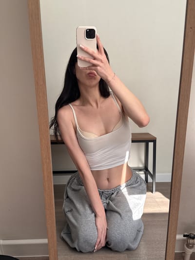 A young woman with long dark hair is kneeling on the floor, holding a smartphone in her right hand to take a mirror selfie. She is wearing a white crop top and grey sweatpants, and has a belly button piercing. The background includes a large mirror with a wooden frame, a wall, and a small table with a metal frame and wooden top. Sunlight streams in from the right, casting shadows on the floor. The scene appears to be indoors, possibly in a residential setting. There is no visible indication of the city of Zakharovo, Russia.