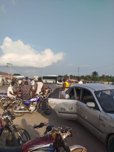 The image depicts a busy street scene in Uduagba, Nigeria. A silver car with significant damage, including scraped paint and dents, is parked with its passenger door open. Several people are gathered around the car and on motorcycles, suggesting a possible incident or traffic congestion.

In the foreground, multiple motorcycles are parked, some with riders. One motorcycle is prominent, displaying a colorful design on its fuel tank. Behind the car, a white bus is visible on the road, along with other vehicles, including trucks. The background shows buildings, shops with awnings, and lush green palm trees, indicating a tropical environment. Power lines and poles are also present, typical of an urban or semi-urban setting.

The sky is partly cloudy with patches of blue, suggesting it's daytime, likely late afternoon given the warm lighting and shadows. The atmosphere appears to be that of a typical day in a bustling Nigerian town, with people and vehicles moving about. The open door and damaged car hint at a potential traffic accident or a breakdown, prompting onlookers to gather. The overall mood is one of everyday activity and movement. No visible text is discernible in the image.