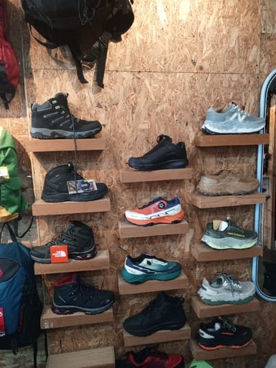 This image captures an indoor retail display showcasing a variety of outdoor footwear, likely in a store in Baza, Myanmar. The setting features a rustic wall made of textured oriented strand board (OSB) or plywood, against which multiple pairs of shoes are arranged on small, individual wooden shelves.
The display features both hiking boots and trail running shoes. On the left side, several pairs of hiking boots are visible, mostly in dark tones like black, dark grey, and olive green. One black mid-cut boot has a tag clearly displaying "GORE-TEX," indicating its waterproof features. Another dark-colored hiking boot below it has a red tag from "THE NORTH FACE."
On the right side, various trail running shoes are displayed. These include several pairs from "New Balance," distinguishable by their logo. One notable New Balance pair is light grey with a thick, textured white sole, while another is dark green with a similar robust sole. Other shoes include a vibrant orange and white pair, a teal and white pair, a solid black athletic shoe, and a black and orange pair, all designed for outdoor terrain.
Above the shoe display, a black backpack hangs from the wall, and parts of a green and blue backpack are visible in the lower left corner, further indicating an outdoor gear store. No people are present, and no specific activity is taking place other than the static presentation of merchandise. The lighting suggests an indoor environment, making the time of day or weather undeterminable.
