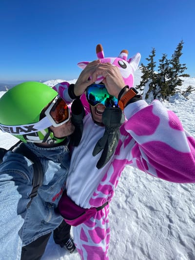 Two individuals are pictured in a close-up, outdoor shot on a snowy mountain slope in Jahorina, Bosnia and Herzegovina, under a clear blue sky. The person on the left wears a bright green ski helmet with white-framed goggles and a blue denim jacket over a backpack, angled towards the second individual. The person on the right wears a white and pink spotted full-body animal onesie, resembling a giraffe or cow, complete with a hooded headpiece featuring ears and small horns. This individual wears blue mirrored ski goggles, black gloves, an orange wristband, and a pink fanny pack, with an open-mouthed expression suggesting excitement. Their gloved hands are raised above their head. In the background, a snowy landscape with several snow-covered evergreen trees and distant snow-capped mountains is visible under bright sunlight.