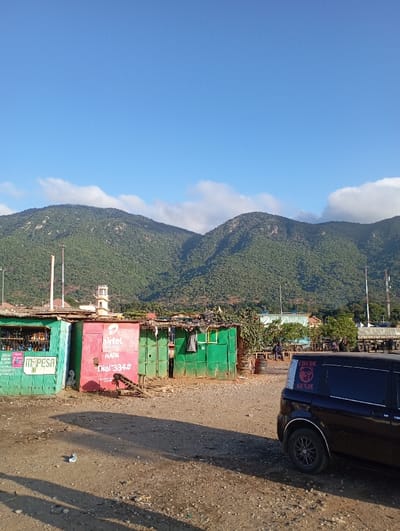 The image presents a sunlit scene in Namanga, Kenya, featuring a rural or semi-urban setting at the base of lush, green mountains. A clear, bright blue sky with a few wispy white clouds stretches above the landscape.
In the midground, a row of informal structures, likely kiosks or small shops, are prominently displayed. The leftmost building is green and features the "M-PESA" logo, indicating a mobile money service. Adjacent to it is a red and green structure with "airtel Money HAPA" and "Dial 334#" visible, suggesting another mobile banking point. Further to the right are more green corrugated metal buildings and makeshift shelters, some with clothes hanging outside, hinting at habitation or small businesses. A tall, white, multi-tiered structure, possibly a mosque minaret or tower, is visible behind these buildings. Utility poles and masts are scattered throughout the background.
The foreground consists of a wide expanse of uneven, light brown dirt and gravel, with shadows cast across it. To the right, a dark-colored van or SUV is parked, its rear visible. The back window of the vehicle displays a white graphic featuring a roaring animal, possibly a lion or tiger, above the text "CHUDE GENJE". In the far distance, near some other structures, a few indistinct figures can be seen, indicating human activity. The overall impression is a vibrant roadside or market area, bustling with daily life under the watchful presence of the towering, forested mountains. The bright lighting suggests it is daytime, possibly morning or late afternoon.