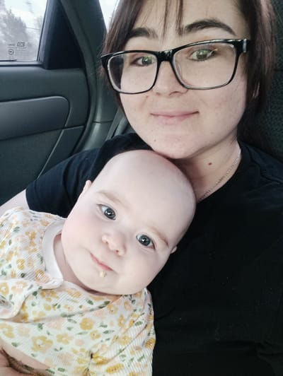 The image presents a close-up portrait of a young woman and an infant, likely taken as a selfie inside a car in Simakovo, Russia. The woman, positioned on the right, appears to be in her 20s or early 30s, with dark hair, bangs, and dark-rimmed glasses. She has fair skin with freckles and sports a gentle, closed-mouth smile, looking directly at the camera. She wears a black t-shirt and a thin silver chain necklace.
Resting on the woman's shoulder and chest, the infant is on the left side of the frame, gazing intently at the camera with wide blue eyes and a curious, neutral expression. The baby has fair skin, light eyebrows, and a small, light-colored crumb or piece of food on its lower lip. The infant is dressed in a light-colored, long-sleeved top adorned with a delicate floral pattern featuring yellow, peach, and green elements.
The background reveals the interior of a car, with a dark gray door panel visible on the left. Through the car window in the upper left, a blurred outdoor scene, possibly showing trees or an indistinct landscape, is visible under natural daylight. Mirrored text can be faintly discerned on the window, with characters resembling "SMDAT" and "KODAK" or similar branding, although much of it is unreadable due to reflections and blur. The overall mood conveys warmth and affection between the two subjects.