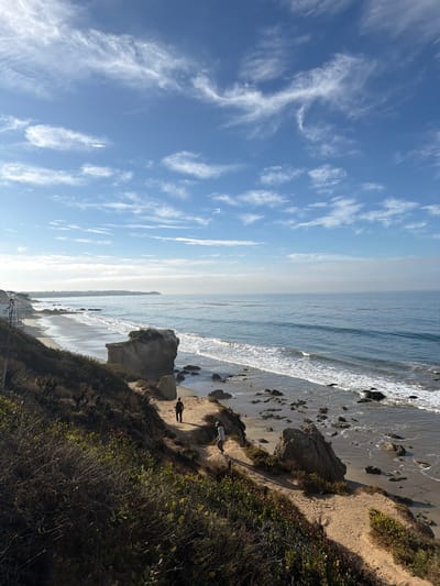 A vertical, medium shot captures a coastal scene in Malibu, California, bathed in the soft light of a partly cloudy day. The foreground is dominated by a steep, dry, and overgrown hillside covered in brush and foliage, with a sandy path winding down towards the beach. Two individuals are present on the path. One, wearing dark clothing, stands near a large rock. Another person, in lighter clothing, walks further down the path. The middle ground features a sandy beach with scattered rocks and waves breaking on the shore. In the background, the vast Pacific Ocean stretches to the horizon, meeting a hazy sky filled with wispy white clouds. The coastline extends to the left, revealing houses nestled along the cliffs. The overall impression is one of serene natural beauty.