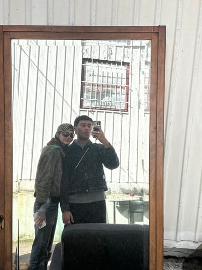 The image displays a mirror selfie of two individuals, a presumed male and a female, in Neuilly-Plaisance, France. The scene is reflected in a vertical mirror featuring a medium-brown wooden frame. The mirror is positioned outdoors, reflecting a weathered white corrugated metal wall as the background. A rectangular window, secured by white horizontal and vertical bars forming a grid, is visible in the reflection behind the subjects.
The individual on the left, presumed female, wears a dark, patterned headscarf, dark sunglasses, a fuzzy olive-green jacket, and blue jeans. She holds a white smartphone in her left hand at her hip. The individual on the right, presumed male, holds a dark-colored smartphone with a visible multi-lens camera array in his right hand, extended forward to capture the reflection. He wears a dark jacket over a light grey sweater and dark trousers. A thin silver chain strap crosses his torso, and he wears a ring on his left ring finger. His lips are slightly puckered. Both individuals stand in close proximity to each other.
The environment suggests an outdoor setting under bright natural light, evidenced by shadows cast on the corrugated wall. The lower portion of the mirror reflects part of a dark, upholstered object, consistent with a chair, and a paved ground surface with some debris.