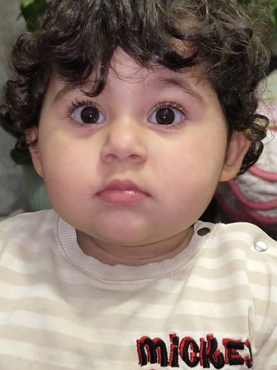 A close-up captures a baby with dark curly hair and big brown eyes. The child's face, featuring a small nose and slightly parted lips, is tilted towards the viewer. The baby is wearing a striped white and beige outfit with "Mickey" embroidered in red and black on the side. A glimpse of greenery is visible in the bottom left corner.