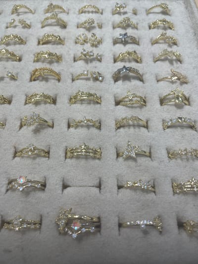 This image shows a top-down view of a display case filled with gold rings. The rings are arranged in rows and columns. Each ring has a unique design, with some featuring small diamonds or other gemstones, while others are plain gold bands. The rings are displayed on a textured, grey background that appears to be velvet or felt. The display case is well-lit, and the rings are in focus, showcasing their intricate details. The image does not contain any information about the city of Yunus Emre, Turkey, nor does it depict any people, actions, or interactions.