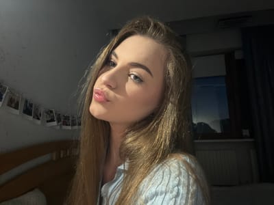 A young Caucasian woman with long, blonde hair is captured in a close-up, low-angle selfie. She is puckering her lips and looking towards the camera. Her eyes are blue and her eyebrows are well-defined. She is wearing a light blue knitted sweater. The background suggests an indoor setting with a bed visible on the left, adorned with a string of small photos. A window with dark curtains is on the right, with a radiator beneath it. The overall lighting is dim, typical of an indoor evening or nighttime scene. There are no visual indicators that place this image specifically in Dnipro, Ukraine.