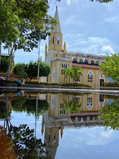 The image depicts a yellow church with a tall, pointed steeple and a clock face, possibly the Iglesia Nuestra Señora de la Soledad in San José de Barlovento, Venezuela. The church is reflected in a shallow body of water in the foreground, which also shows reflections of trees and the sky. There are trees and bushes bordering the church and the water. One person is visible sitting on the edge of the water, facing away from the camera, wearing a blue shirt. Another person is partially visible sitting on a ledge to the left. The sky is blue with scattered white clouds. The overall scene suggests an outdoor public space on a bright, possibly sunny day.