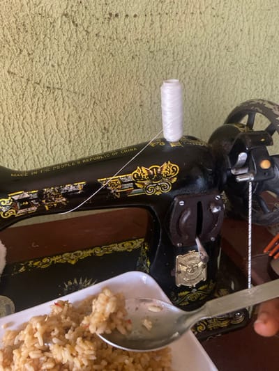 A close-up, slightly overhead shot captures a vintage black sewing machine adorned with gold floral and ornamental patterns. A white spool of thread is mounted on a spindle on the machine's left side, with the thread extending upwards towards the machine's threading mechanism. The sewing machine exhibits signs of wear and tear, with chipped paint revealing the underlying metal. Vertically printed on the machine's arm, the text "MADE IN THE PEOPLE'S REPUBLIC OF CHINA" is clearly legible.
To the right of the sewing machine, a white plate holding a serving of rice is partially visible, with a metallic spoon resting within it. The rice appears to be a cooked dish, possibly fried rice or jollof, with small flecks of red ingredients interspersed throughout. The background is a textured, light green surface, possibly a mat or fabric. The scene suggests a domestic setting in Oba-Aboji, Nigeria, where a meal is present near the sewing equipment. No direct human activity is depicted, and the lighting indicates it is daytime.