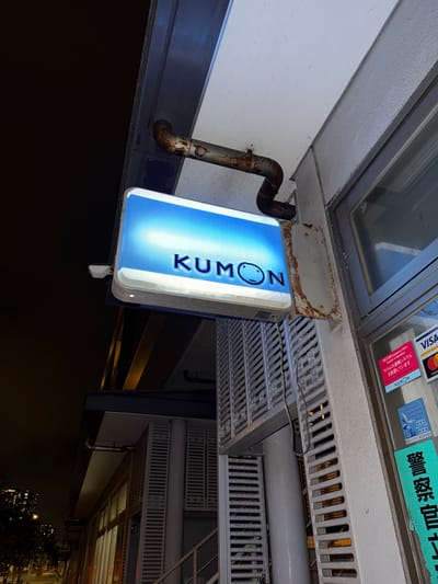 The image shows a nighttime exterior view of a Kumon learning center in Chiba, Japan. A lit Kumon sign with the logo, which includes a smiley face, is prominently displayed. The sign is attached to the building by a rusty metal pipe. Below the sign, a window with a grey grated facade is visible, and to the right, there are stickers on the wall, including one for VISA and Mastercard, a "SECOM Emergency Alarm System" sticker, a sticker for "MARUHARA BAYTOWN SHOPPING MALL," and a green sticker with Japanese characters that appear to be related to police. In the background, city lights and buildings are visible, suggesting an urban environment. The overall scene is focused on the building's entrance and signage.