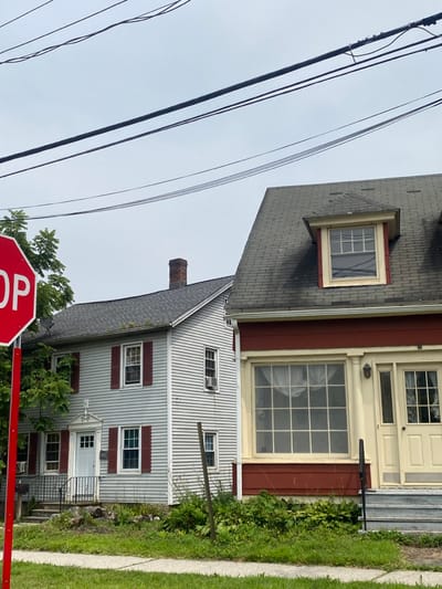 The image displays a street scene in Ridgefield, United States, featuring two residential buildings under an overcast sky. Overhead, multiple dark electrical wires traverse horizontally across the frame. On the left, a red octagonal STOP sign with white lettering is partially visible on a red pole.
The house on the left is a two-story structure with light grey horizontal siding and a dark grey shingled roof exhibiting some moss growth. A brick chimney rises from the roof. The building has multiple windows fitted with red shutters. A white front door is located on the ground floor, accessible via grey stone steps with a black metal railing. An air conditioning unit is installed in a window on the second story, right side of the building.
The house on the right is a two-story building characterized by a dark red facade on the ground floor and a dark grey mansard-style roof. The ground level features a large, multi-paned window with light yellow trim, positioned to the left of a light yellow main entry door. The door leads to a small porch with light-colored steps and a black railing. A dormer window with light yellow trim is present on the roof.
The foreground consists of a concrete sidewalk and green grass with scattered patches of overgrown foliage. A vertical wooden stake is visible between the two houses in the ground. No individuals are present, and no specific actions or interactions are occurring within the scene.