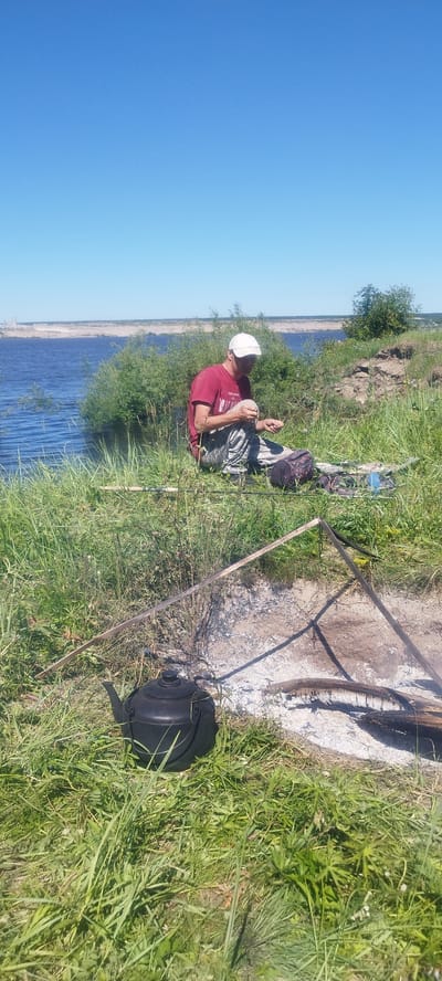 A man in a red shirt and camouflage pants sits on a grassy bank next to a body of water, holding something in his hands. He is wearing a white cap. In front of him are a black kettle and some camping gear. A makeshift fire pit with ashes and burnt wood is also visible. The background shows a wide river or lake, lush green vegetation, and a clear blue sky. The location is identified as Mokhsogollokh, Russia.
