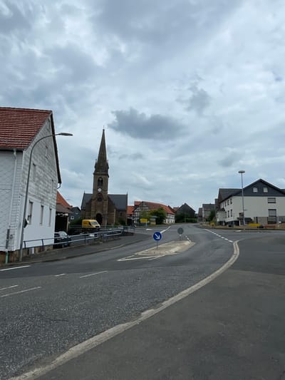 The image depicts a street scene in Seigertshausen, Germany, under a cloudy sky. The photograph is oriented vertically. In the foreground, a white building with a red-tiled roof and a metal fence is visible. To the left of this building, a street lamp stands. Behind this structure, a tall, pointed church spire dominates the middle ground, with a clock face visible on its tower. The church is made of stone and has arched doorways and windows. A yellow van is parked near the church. Further back, several houses and buildings line the street, showcasing a mix of architectural styles, some with modern dark roofs and windows, and others with traditional red tiles. The street itself is paved and curves to the right. A blue traffic sign with a white arrow indicates a mandatory right turn. The overall impression is a quiet, residential area, likely in the afternoon, given the overcast conditions. There is no visible text in the image, other than on the traffic sign.