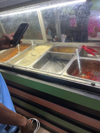 The image displays an indoor scene featuring a glass-covered food service display unit, likely within a cafeteria or restaurant in Isasi, Nigeria. The unit's base is horizontally striped in alternating green, brown, and black.
The foreground is dominated by the left side of a dark-skinned individual, wearing a light blue short-sleeved shirt with a partially visible logo on the sleeve. This person's left hand, positioned at eye level, holds a dark-colored smartphone vertically, with its screen facing away from the viewer. A portion of their right arm and hand is visible lower down, holding a coiled white cable, possibly a charging cable.
Behind the person, the food display unit features multiple stainless steel trays under a glass cover illuminated by a fluorescent strip light. From left to right:
1. A tray containing an orange-red granular food item.
2. A tray filled with a light yellow granular food item, accompanied by a silver serving spoon.
3. A largely empty tray with minimal residue.
4. A tray containing a dark red stew or sauce, with a silver serving ladle and a pair of red-handled tongs.
Reflections on the glass cover show indistinct figures and shapes, indicating other individuals or elements within the immediate vicinity of the food display.