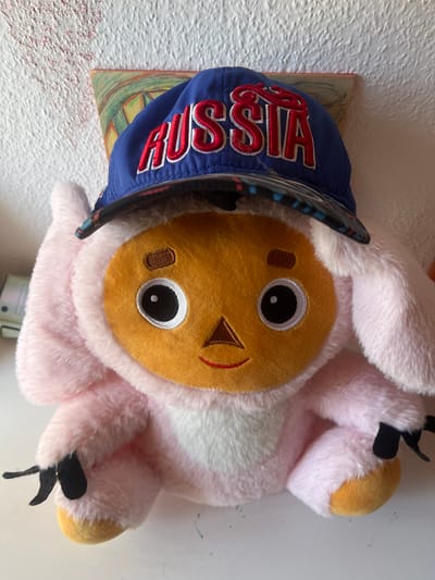 A pink, plush toy with a yellow face, black eyes with white pupils, and a small brown nose and red smile is wearing a blue baseball cap. The cap has "RUSSIA" embroidered in red letters with white trim. The toy's ears are long and pink, and its hands appear to have black claws or fingers. It is sitting on a white surface. In the background, a textured white wall is visible, with a small, abstract painting or artwork hanging slightly off-center behind the toy. The overall scene appears to be indoors, possibly in a domestic setting, but there is no direct visual evidence to confirm its specific location within Schwäbisch Hall, Germany.
