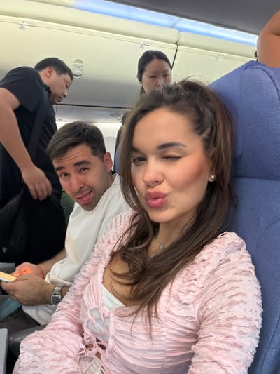 Description of the image:

Location: Inside an airplane cabin; uncertain if near Shunyi District, China.

People:
* A woman with long brown hair is positioned in the foreground. She is winking and puckering her lips. She is wearing a pink top with textured ruffles and a necklace.
* A man behind her is sticking his tongue out playfully. He is wearing a white shirt.
* Two other individuals are visible in the background, appearing to be seated in the airplane. One is wearing a black shirt.

Objects:
* Airplane seats.
* A blue headrest.
* The woman has a wristwatch on her left wrist.
* An airplane window.
* Overhead compartments are visible along the side of the airplane.

Actions:
* The woman is posing for the photo.
* The man behind her is making a face.
*The others look like they are relaxing or sleeping.

Interactions: The people appear to be traveling together and taking a photo inside the airplane.

Environment:
* The interior of an airplane, including the overhead compartments, seats, and window.