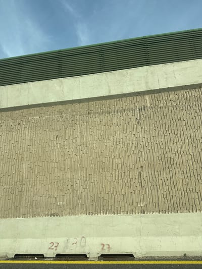 The image displays a concrete wall structure, likely part of a sound barrier or retaining wall, situated in Kfar Shmaryahu, Israel, under a clear light-blue sky with minimal cloud cover. The upper section of the structure features a dark green, horizontally corrugated barrier mounted above a smooth, light beige concrete band. Below this, the primary feature is a large expanse of light brown concrete textured with a repeating pattern of vertical, staggered rectangular reliefs. At the base of this textured wall, another smooth, light beige concrete section is visible. On this lower concrete section, near ground level, three numbers are painted in red: "27", "30", and "27". Immediately below these markings, three rectangular metal drainage grates are set into the concrete. The very bottom of the image shows a solid yellow line on a dark road surface, indicating the edge of a paved roadway. No individuals or active movements are present.