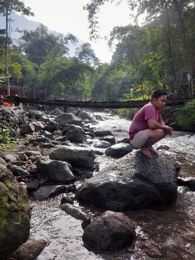 A man in a pink shirt and light shorts sits cross-legged on a large, dark rock in a shallow river. He faces away from the camera, looking down the river. The water flows around and between many smaller rocks. Lush green trees line the riverbanks. A wooden bridge, with people standing on it, spans the river in the background. Sunlight streams through the trees.