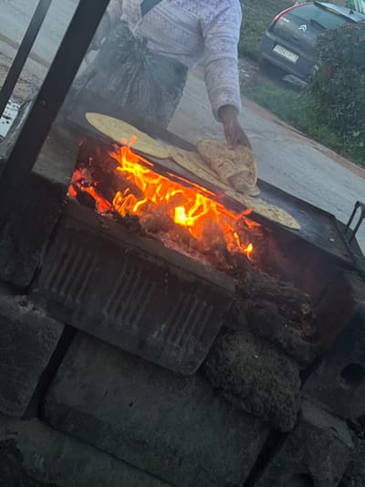 The image captures an outdoor cooking scene, likely during dusk or early evening given the prominent glow of the fire. The photograph is taken from a slightly elevated and tilted perspective, looking down into a large, rectangular outdoor cooking setup.

In the center of the frame, bright orange and yellow flames erupt from a long, narrow fire pit. Above the fire, a large, dark metal griddle or "tava" extends, on which several round, flatbreads are being cooked. One person is visible on the left side of the frame, their arm and torso in focus. They are wearing a light-colored, possibly lavender or white, long-sleeved garment with a patterned design, and a dark patterned skirt or lower garment. Their hand is holding up one of the flatbreads, appearing to be in the process of placing it on or turning it on the hot griddle. Another flatbread is clearly visible laid flat on the griddle closer to the flames, and others are partially visible.

The cooking structure itself appears to be a makeshift setup, constructed from various dark-colored bricks and rough stones. One distinct brick on the right side of the fire pit has a ribbed texture. On the far side of the griddle, a metal tool, possibly tongs or a scraper, can be seen resting.

In the upper left background, a dark-colored hatchback car, possibly a Citroën given the emblem visible, is parked on what appears to be a paved or dirt road. Part of its license plate is visible, showing "51914 A". Behind the car, green foliage suggests the presence of bushes or trees. The ground immediately surrounding the cooking area looks unpaved. The overall impression is one of traditional outdoor cooking.