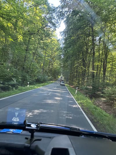 The image captures a view from inside a vehicle traveling on a two-lane road bordered by a dense forest. The road appears to be in good condition with visible white lane markings and a white shoulder line. Sunlight filters through the canopy of the trees, creating dappled shadows on the road surface. Several trees line both sides of the road, with lush green foliage. In the distance, two vehicles are visible on the road: a silver car and a truck. One vehicle marker post is visible on the right side of the road. The foreground shows the dashboard of the vehicle, with a phone mount and a sticker affixed to the windshield. The overall environment suggests a scenic drive through a wooded area. The image is identified as being taken in Volpertshausen, Germany.