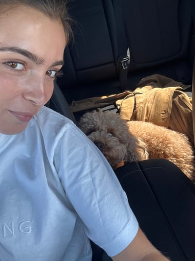 The image depicts the interior of a vehicle, featuring a female and a small dog. A light-skinned female, with dark hair and wearing a light-colored short-sleeved t-shirt with partially visible embroidered text "NG," occupies the left portion of the frame. She looks directly towards the camera, indicating a self-portrait. To her right, a medium-brown, curly-haired dog rests on a dark car seat, its head turned towards the viewer. In the background, dark car seats are visible, along with a tan-colored jacket, appearing to be suede, draped over a rear seat. A silver seatbelt buckle is observable on the upper left rear seat. The image contains no discernible information that identifies its location as being in the city of Pombal, Portugal.
