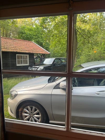 The image, taken from inside a building looking through a window with multiple panes, shows an outdoor scene in Collendoorn, Netherlands. In the foreground, the hood and wheel of a silver car are visible. Behind this, a black Mercedes-Benz SUV is parked. To the left, a dark wooden building with a terracotta tile roof and a white-framed window is present. A person wearing a striped shirt is standing near the black SUV. The background consists of lush green trees. The lighting suggests it is daytime, possibly late afternoon or early evening.